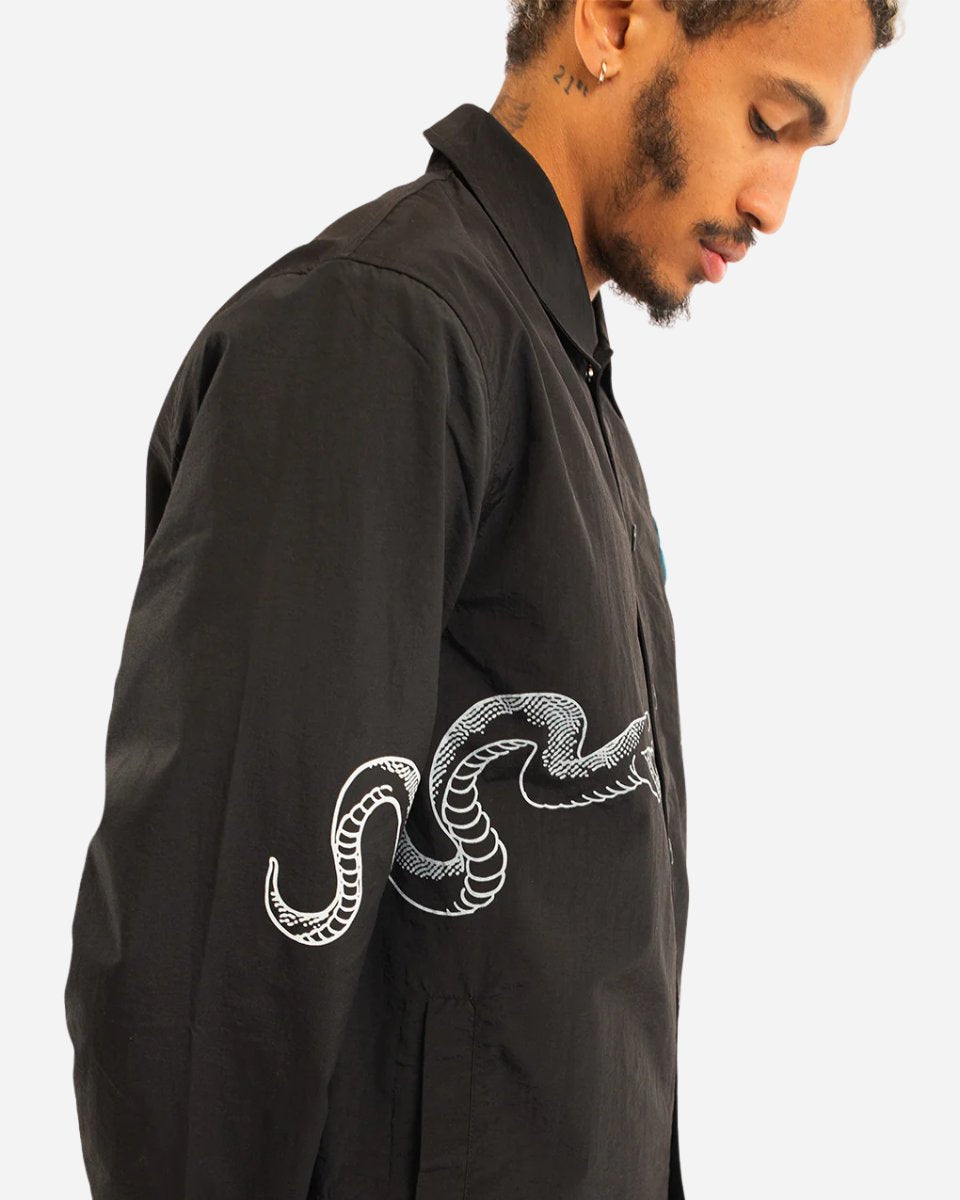 A person wearing the 2025 P&Co Coach Jacket, a black button-up featuring a white snake design wrapping around the sleeve, looks down against a plain light background.