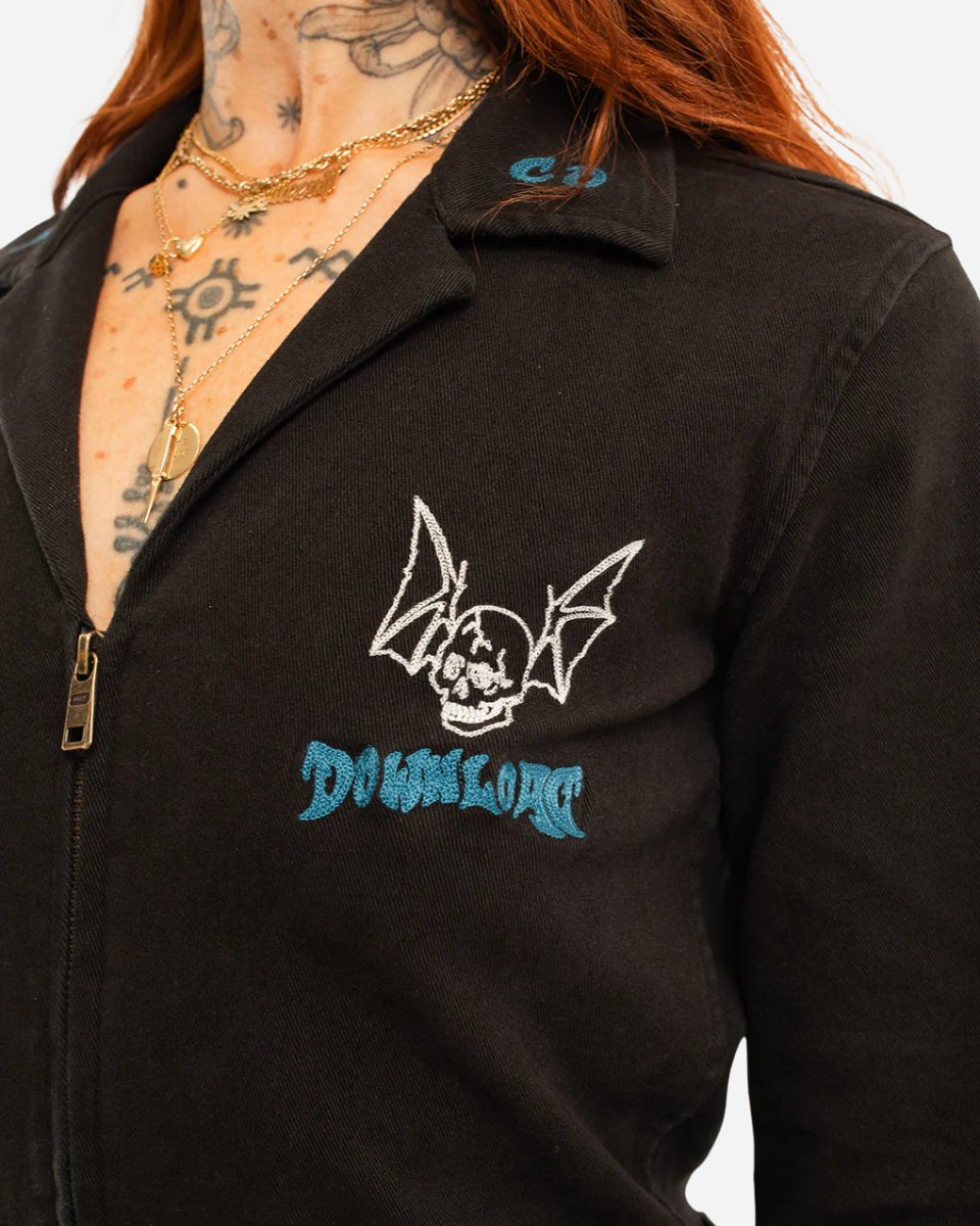 A person wearing the P&CO Long Sleeve Boiler Suit by 2025, featuring a bat-winged skull patch and blue DOWNLOAD text on the chest, with visible chest and neck tattoos and layered gold necklaces.