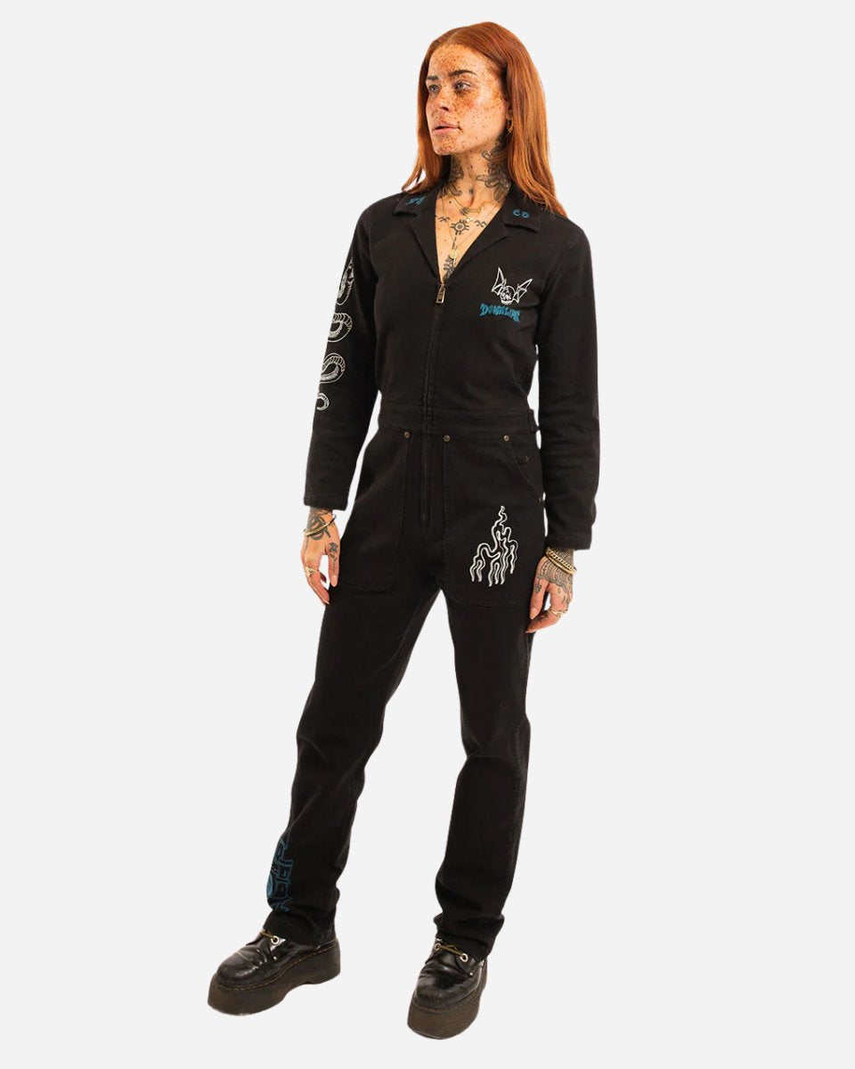 A person with long reddish hair and tattoos wears the 2025 P&CO Long Sleeve Boiler Suit and black platform shoes, standing against a plain white background.