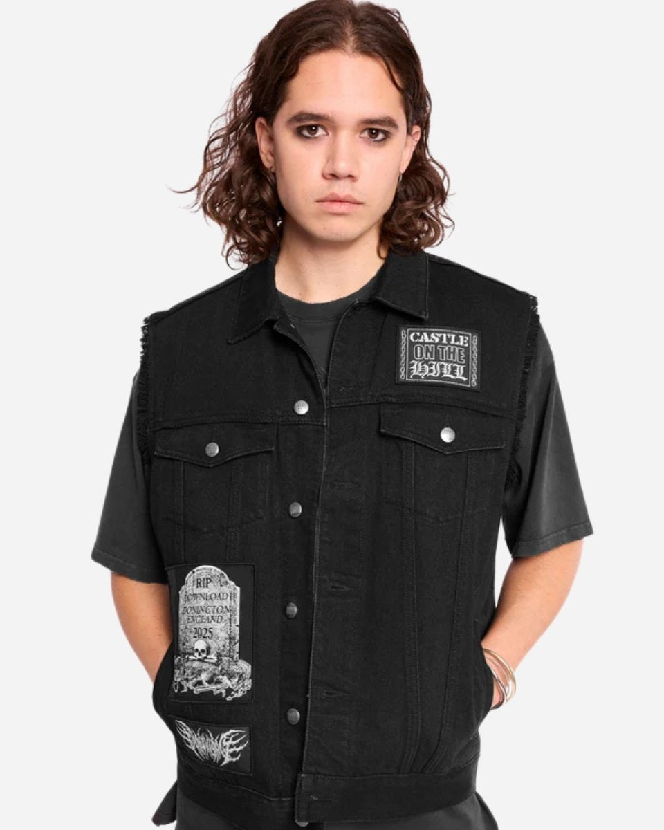 A young person with wavy brown hair wears the Mary Wyatt Patch Lord Battle Jacket by 2025—black denim with silver buttons and gothic patches—over a black t-shirt, standing against a plain light background.