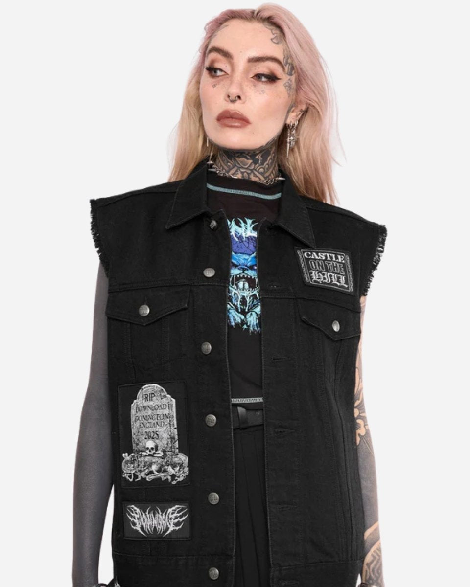 A person with long, light pink hair and tattoos wears the Mary Wyatt Patch Lord Battle Jacket by 2025 over a black shirt with a blue graphic. They have bold makeup, multiple ear piercings, and are looking slightly to the side.