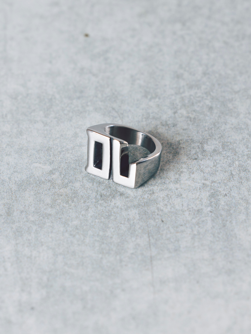 A chunky silver ring resting on a grey surface, featuring the initials "DL" in a bold, block-style design.