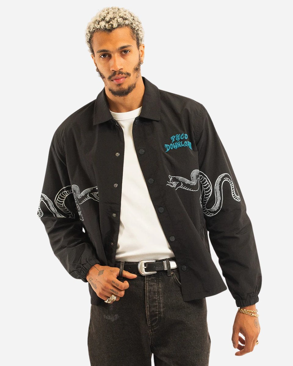 A man with short, curly blonde hair and a beard wears the 2025 P&Co Coach Jacket, black with a white snake graphic on the sleeves and blue PSCO DONSLOR text, paired with a white t-shirt, dark pants, and a belt.