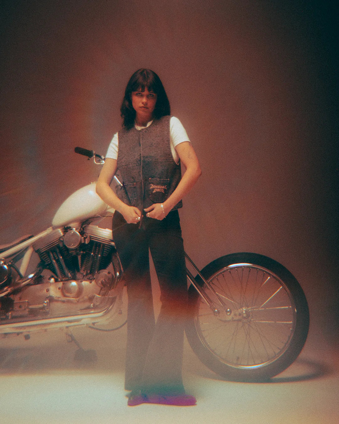 In a softly lit, vintage-inspired studio, a person with shoulder-length dark hair stands before a white motorcycle, wearing the Download 2026 P&Co Nomad Vest over a white tee and dark pants.