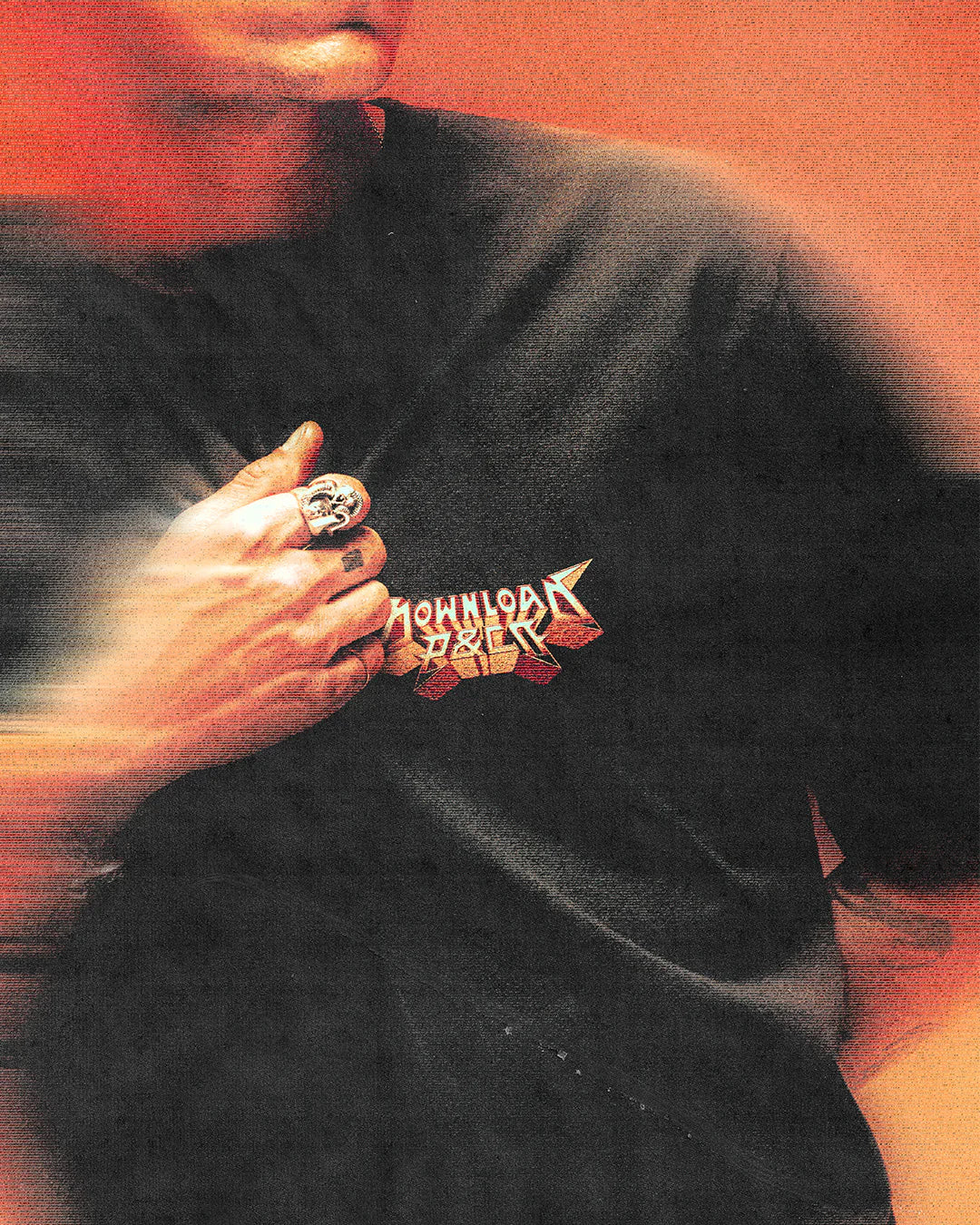 Someone wearing the P&Co Unisex Cobra Tee by Download 2026, featuring colorful graphic text, holds the shirt near their chest while sporting a large ring. The image has a blurred, orange-toned effect.