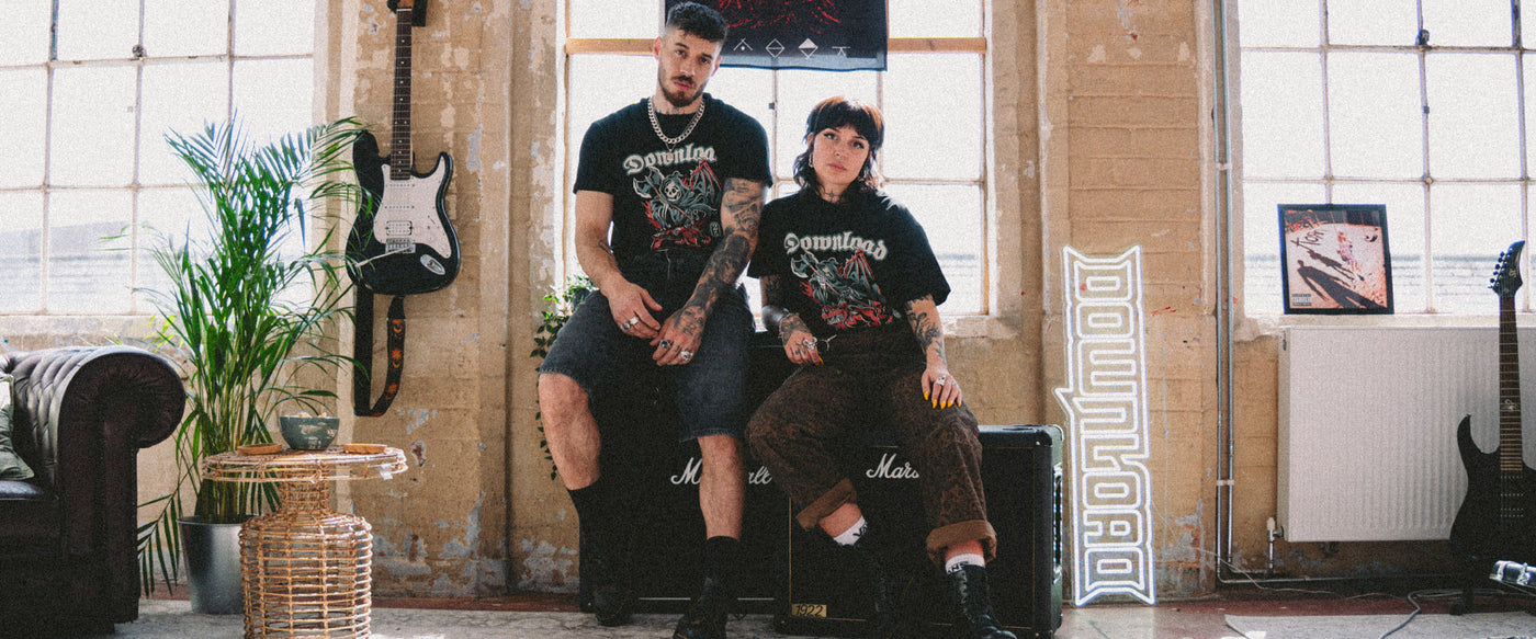 A man and a woman in band t-shirts sit on black Marshall amps in a sunlit room with large windows, guitars, plants, and a neon “DOWNLOAD” sign. The atmosphere is casual and artistic.
