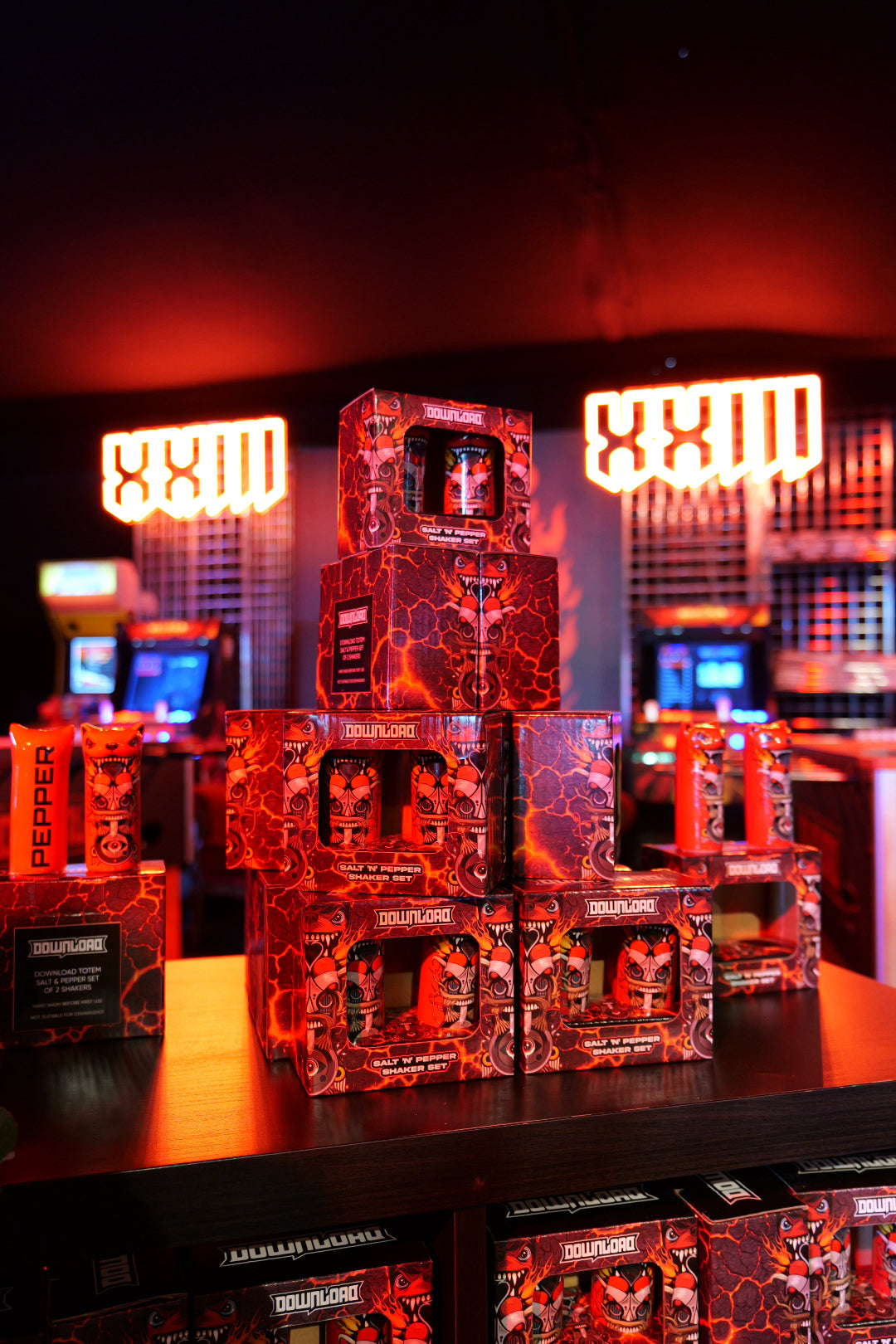 A pyramid display of Download 2026 Totem Salt and Pepper Shakers in red-and-black boxes sits on a counter, with glowing red neon XXIII signs and arcade machines in the background.