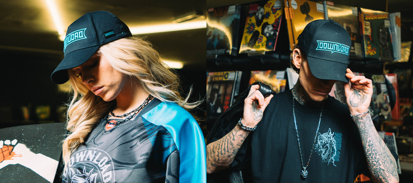 Two people wearing black Download caps and streetwear pose in a comic book shop. The left person has long blonde hair, while the right person has tattoos and lifts the cap brim. Colorful comic books are visible in the background.