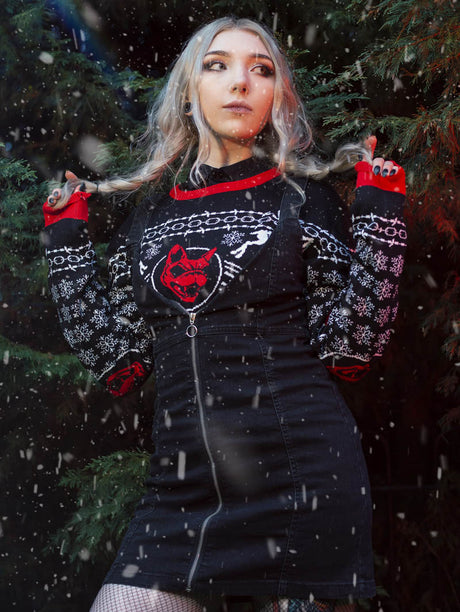 A person with long, wavy blonde hair stands in front of pine trees wearing a festive black jumper with red and white patterns, including the Download Festival dog logo, snowflakes, and skeleton reindeer. The jumper is layered under a black zip-up pinafore dress. Snow is gently falling around them.