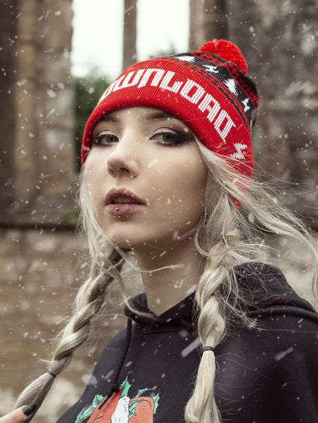 A woman with platinum blonde hair in braids wears a red and black knit Download Festival beanie with white tree and lightning bolt patterns, and a red pom-pom on top. Snow falls around her as she looks into the camera, with a blurred stone background behind her.
