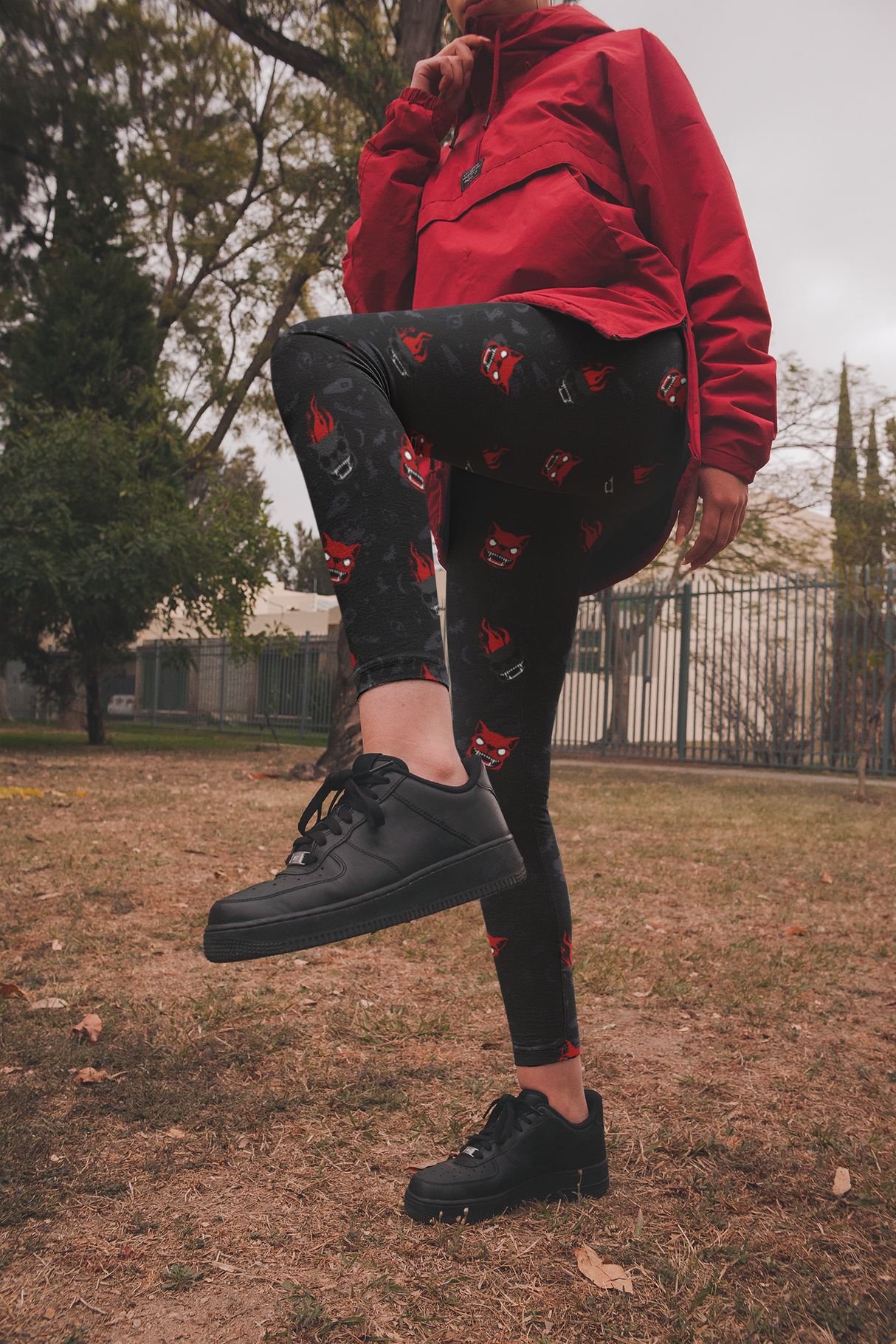 A person outdoors wearing Halloween-themed black leggings decorated with red and grey stylised dog faces and subtle spooky motifs. They are also wearing a red jacket and black trainers, with one leg raised in a dynamic pose against a background of grass, trees, and fencing.
