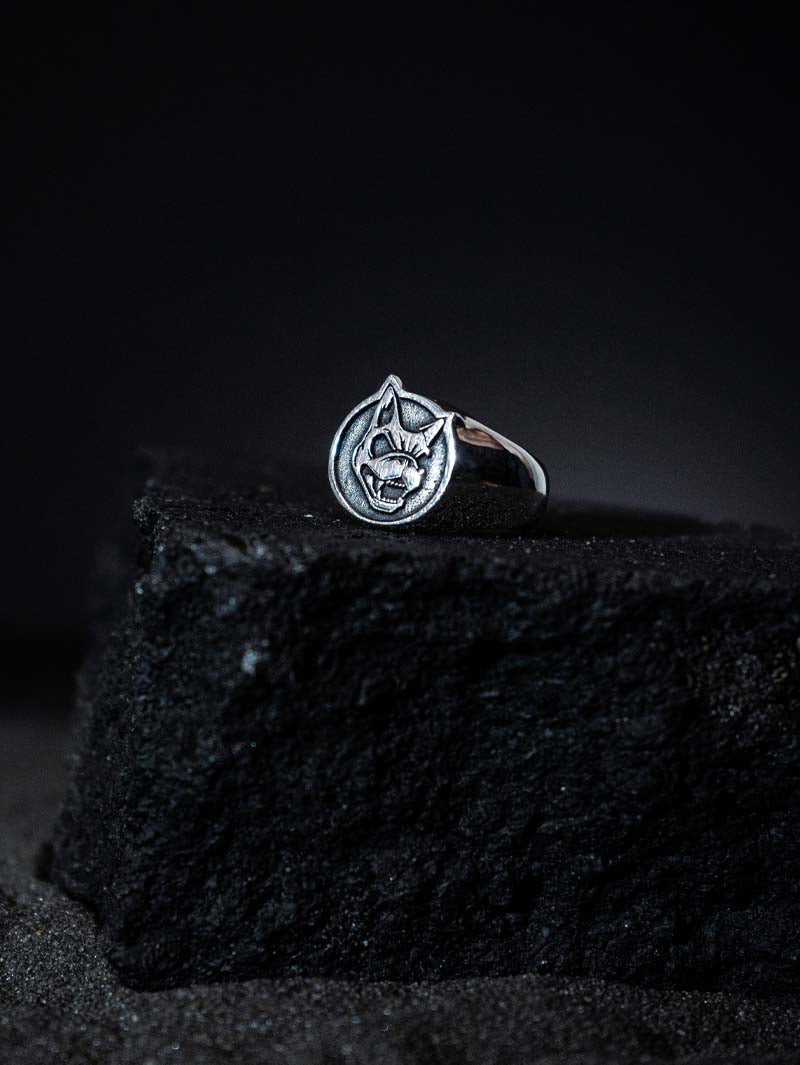 Sterling silver ring featuring an engraved snarling dog head emblem, displayed on a textured black stone block with a dark gradient background.