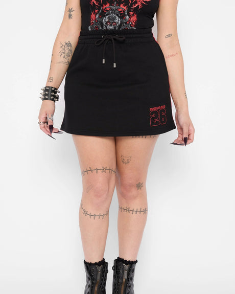 Wearing the Mary Wyatt Defender Sweatskirt by Download 2026, paired with black boots, a spiked bracelet, black top with red graphics; arms and legs feature tattoos and nails are painted black.