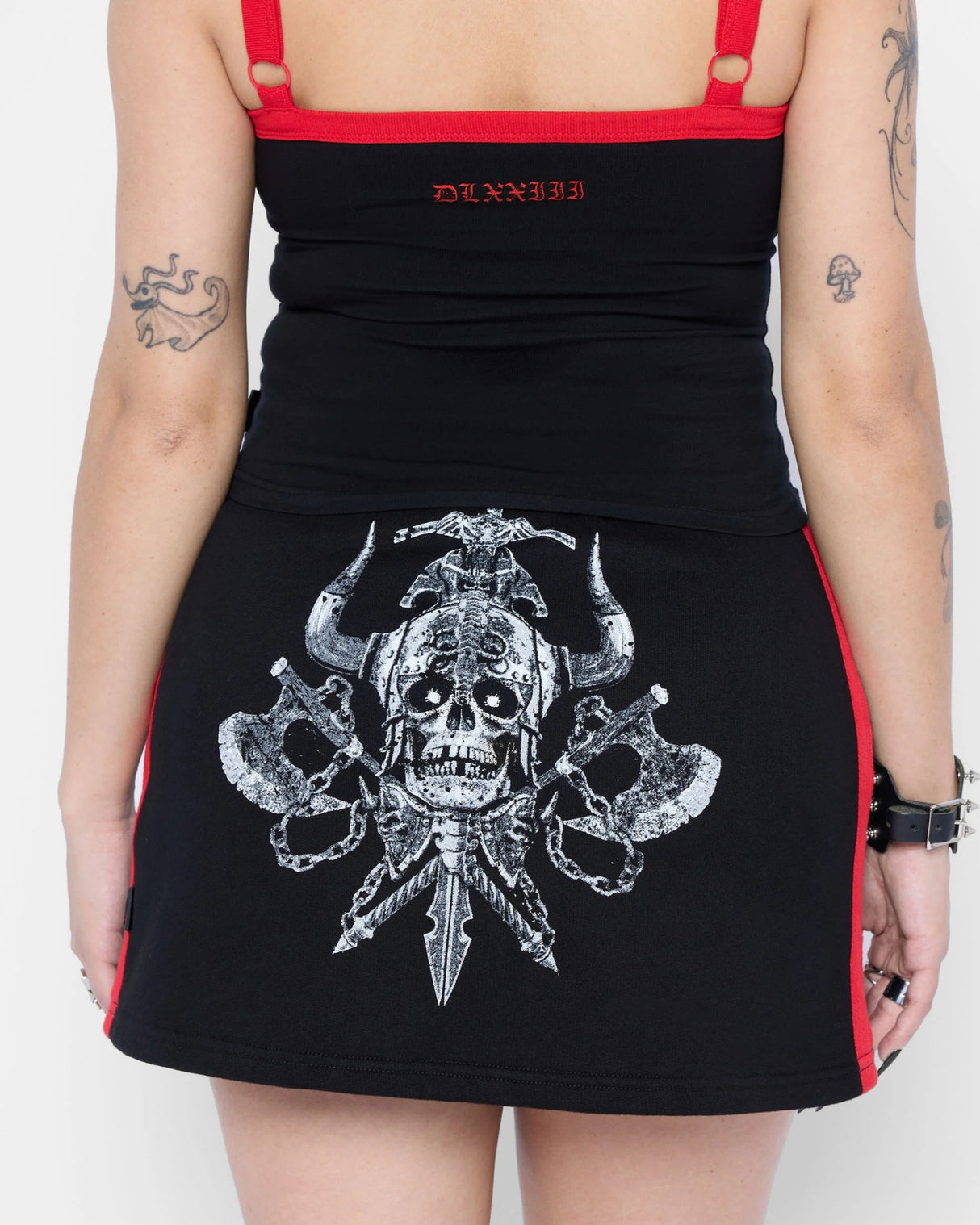 A tattooed person, seen from the back, wears a black Mary Wyatt Defender Sweatskirt by Download 2026 with red accents, a large horned skull graphic with crossed swords and axes, a matching tank top, and a studded bracelet.