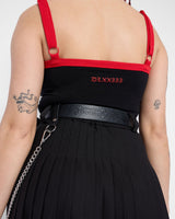 A person wearing a black pleated skirt, a black top with red straps, and the Mary Wyatt Chain Belt by Download 2026. Tattoos are visible on their arm and back. The scene is shown from behind, focusing on clothing and accessories.