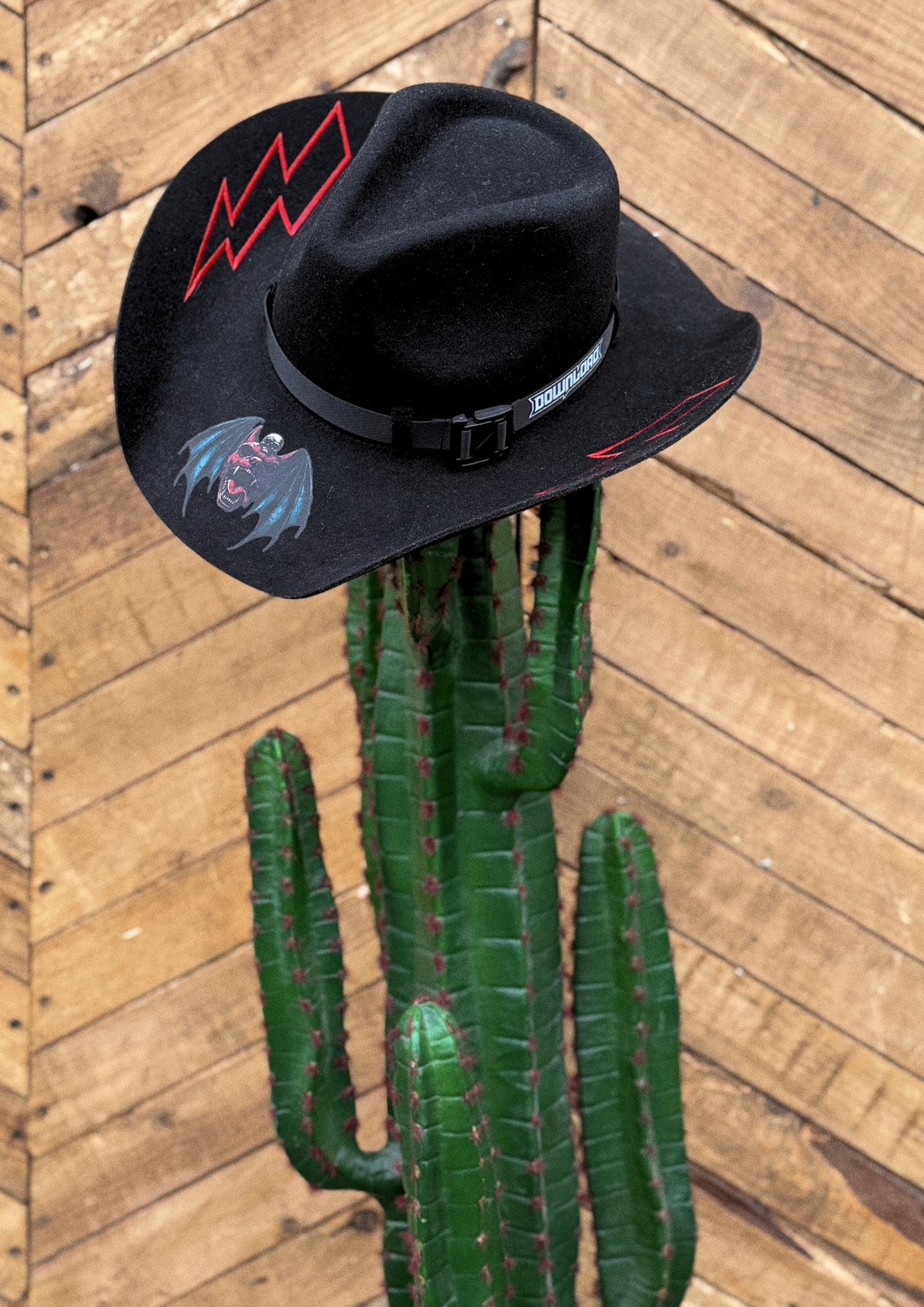 The Download Cowboy Hat by Download 2026, featuring red lightning accents and a painted bat, sits on a tall green cactus with a wooden plank wall background.