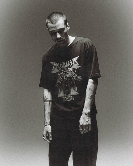 A man with short hair and tattoos stands against a plain background, looking down with a serious expression. He wears the P&Co Line Up T Shirt 2026 by 2026, rings on his fingers, and dark pants, with his arms at his sides.