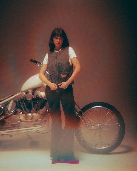 In a softly lit, vintage-inspired studio, a person with shoulder-length dark hair stands before a white motorcycle, wearing the Download 2026 P&Co Nomad Vest over a white tee and dark pants.