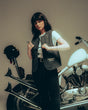 A woman with dark hair stands by a white motorcycle, wearing the P&Co Nomad Vest by Download 2026 over a white tee and black pants. She holds her vest and faces the camera; a black helmet hangs from the bike’s handlebars.