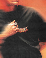 Someone wearing the P&Co Unisex Cobra Tee by Download 2026, featuring colorful graphic text, holds the shirt near their chest while sporting a large ring. The image has a blurred, orange-toned effect.