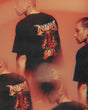 A person wearing the Download 2026 P&Co Unisex Cobra Tee, featuring a colorful graphic and brand name on the back, appears multiple times in a blurred, overlapping reddish-orange effect.