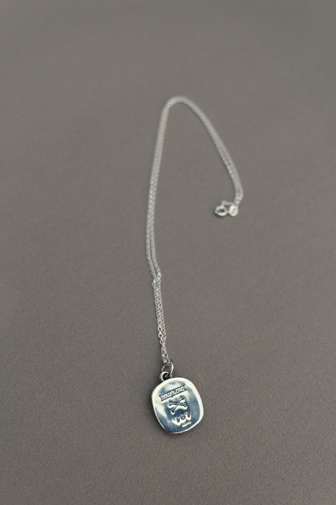 The TGF x Download Necklace by Download 2026 features a thin silver chain and a square pendant engraved with an octopus and the word "HYPNOSIS," shown resting on a gray surface.