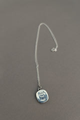 The TGF x Download Necklace by Download 2026 features a thin silver chain and a square pendant engraved with an octopus and the word "HYPNOSIS," shown resting on a gray surface.