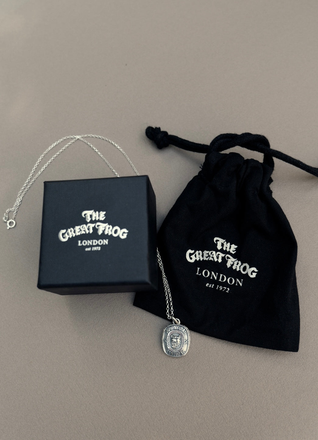 The TGF x Download Necklace by Download 2026, a silver pendant necklace, comes with a black box and drawstring pouch—each featuring The Great Frog London logo—displayed on a light beige surface.