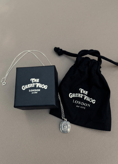 The TGF x Download Necklace by Download 2026, a silver pendant necklace, comes with a black box and drawstring pouch—each featuring The Great Frog London logo—displayed on a light beige surface.
