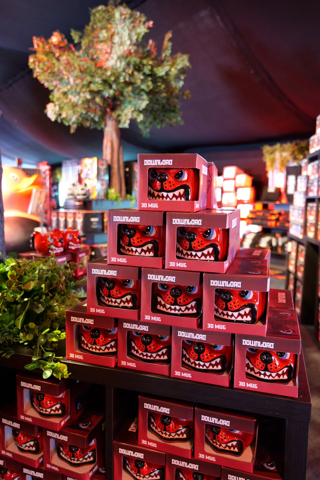 A display of stacked red Dog Head Shaped Mugs by Download 2026, featuring cartoon monster faces, set on black shelves in a shop with artificial plants and trees in the background.