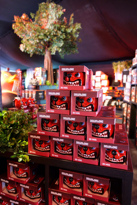 A display of stacked red Dog Head Shaped Mugs by Download 2026, featuring cartoon monster faces, set on black shelves in a shop with artificial plants and trees in the background.